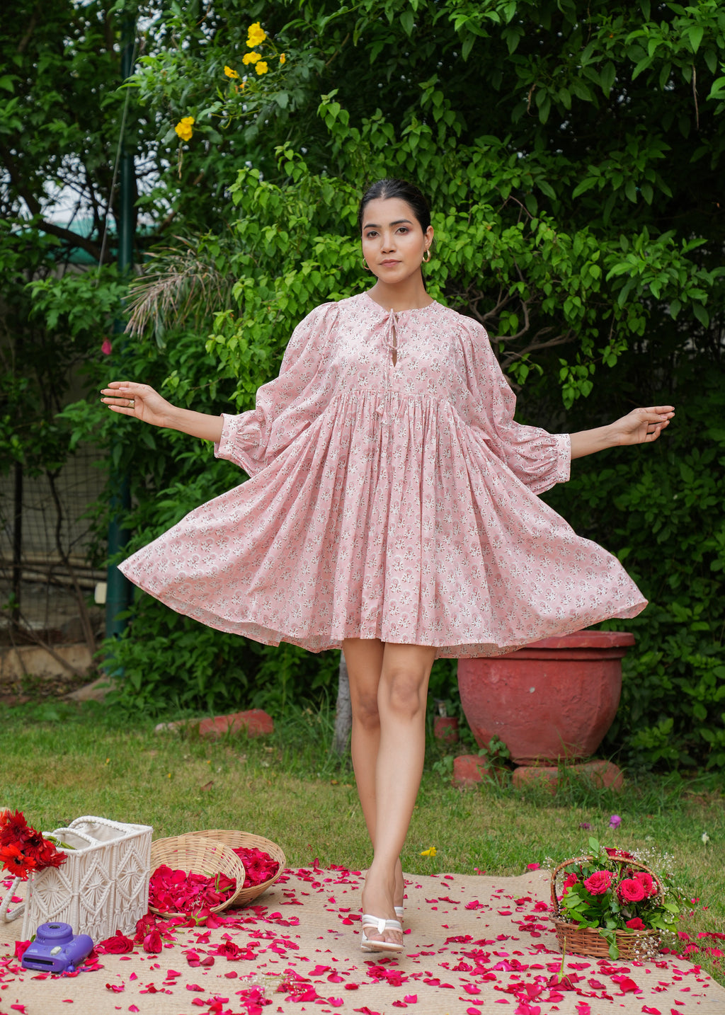 A woman standing on a platform with a scattering of pink flower petals around her, wearing a pink hand-block printed, short-sleeved mini dress.